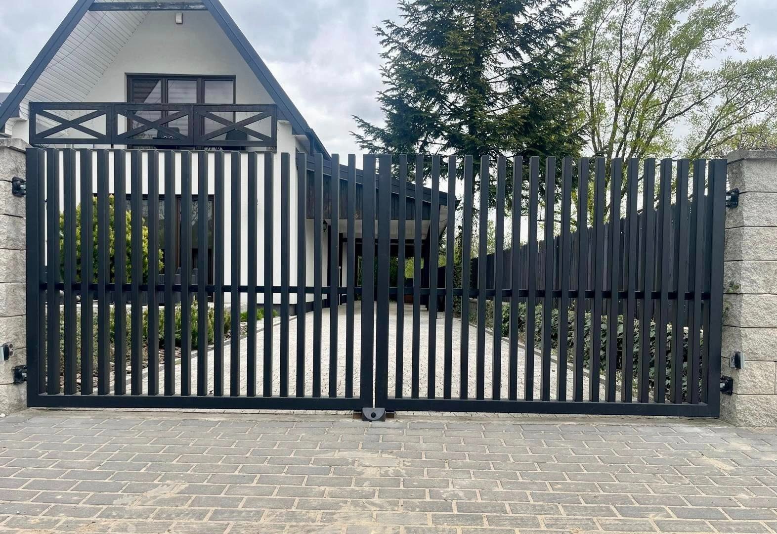 Nowoczesna, czarna brama wjazdowa z pionowych profili, na tle domu z balkonem i brukowanej nawierzchni. Minimalistyczny design, symetryczny układ.