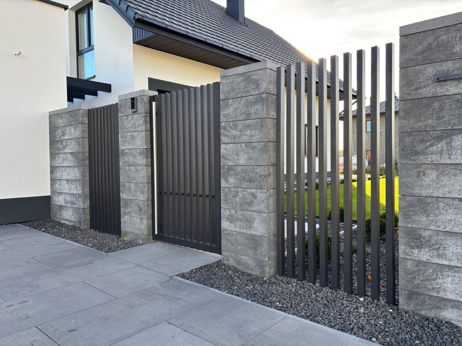 Nowoczesne ogrodzenie panelowe z betonowymi słupkami i metalową bramką w kolorze antracytowym, na tle domu z szarą kostką brukową i kamieniami ozdobnymi.