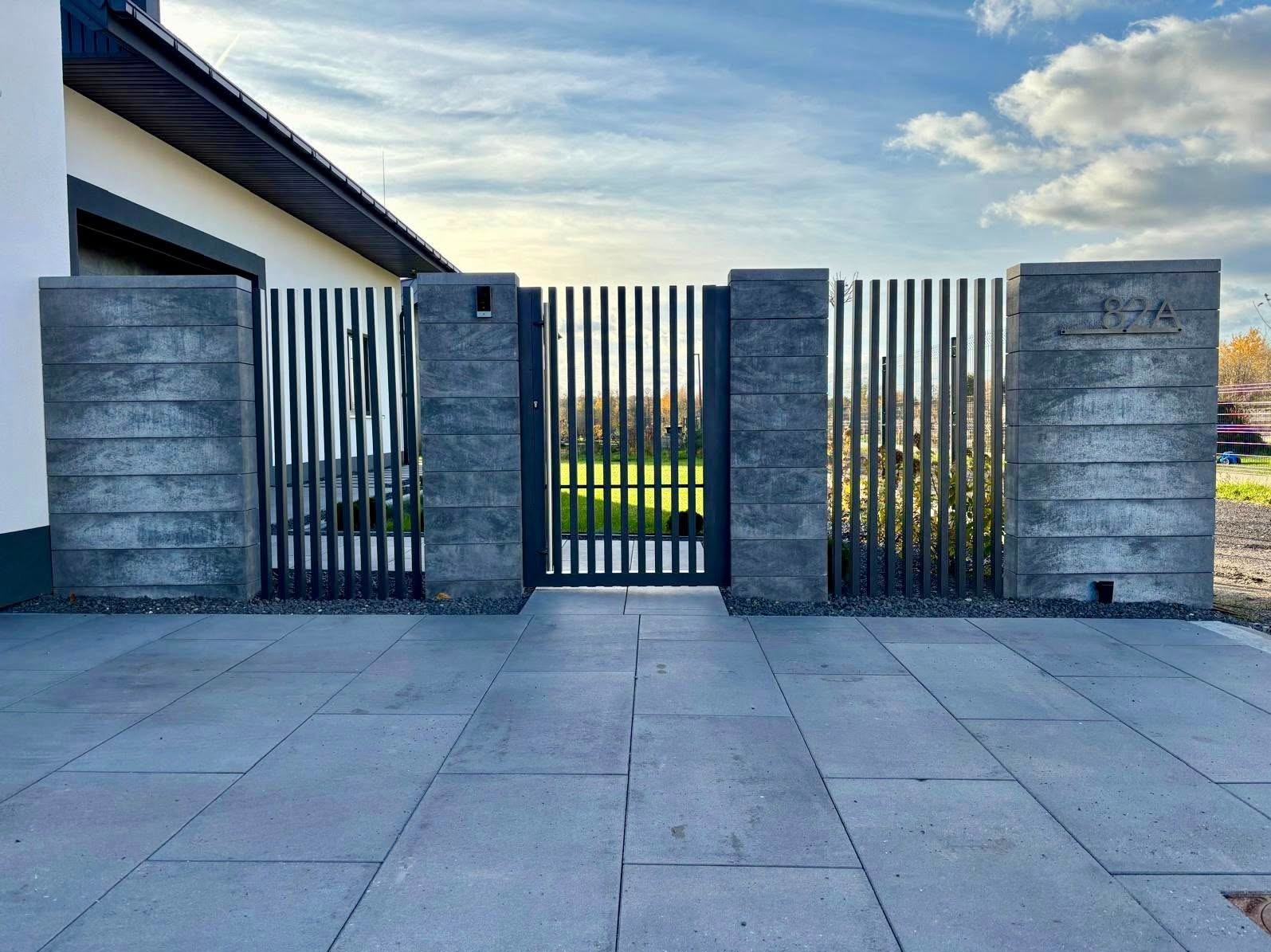 Nowoczesna brama wjazdowa z pionowych profili 60x40, osadzona między betonowymi słupkami z numerem domu 82A. Szara kostka brukowa prowadzi do wejścia.