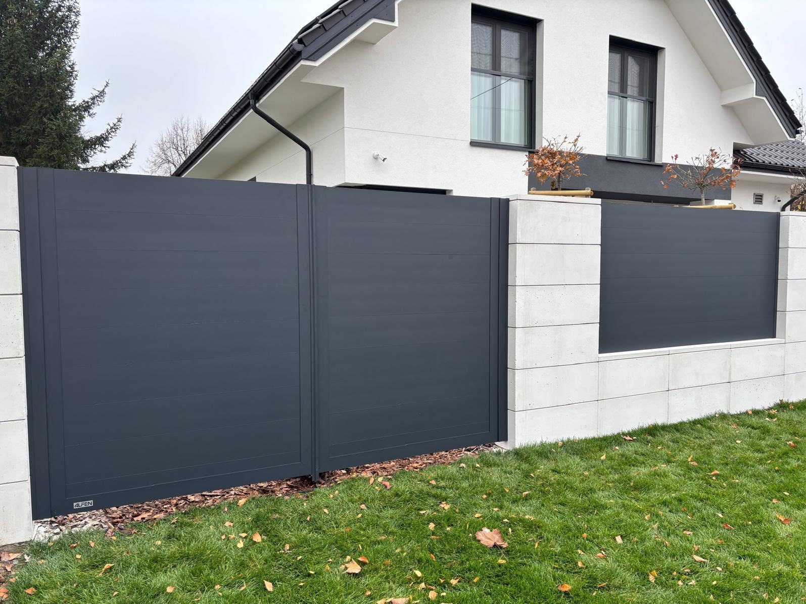 Nowoczesna, przesuwna brama w kolorze antracytu, z poziomymi panelami, wkomponowana w betonowy mur. Minimalistyczny design, trawnik z opadłymi liśćmi.