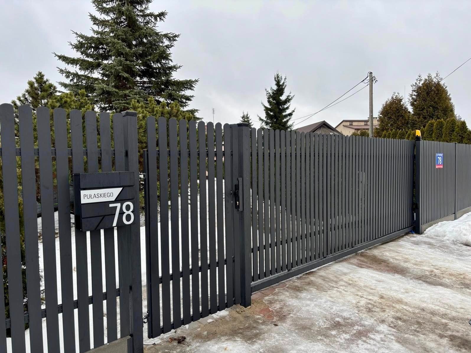 Grafitowa brama przesuwna z furtką i skrzynką na listy z numerem 78, na tle zimowego krajobrazu z iglakami. Nowoczesny design, pionowe sztachety.