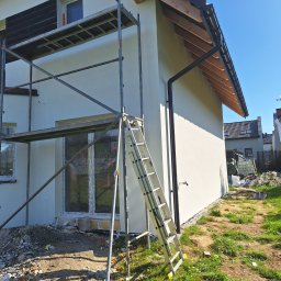 LUISBUD - Ocieplona elewacja domu jednorodzinnego w trakcie prac wykończeniowych, widoczne rusztowanie i drabina. Okna zabezpieczone folią.