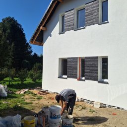 LUISBUD - Praca przy elewacji domu: mężczyzna miesza zaprawę obok białej ściany z drewnopodobnymi panelami. Widoczne wiaderka z materiałami i narzędzia.