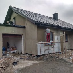 LUISBUD - Dwóch pracowników na rusztowaniu i dachu ociepla elewację domu jednorodzinnego. Widoczne narzędzia i materiały budowlane. Dom z szarą dachówką i oknami zabezpieczonymi folią.