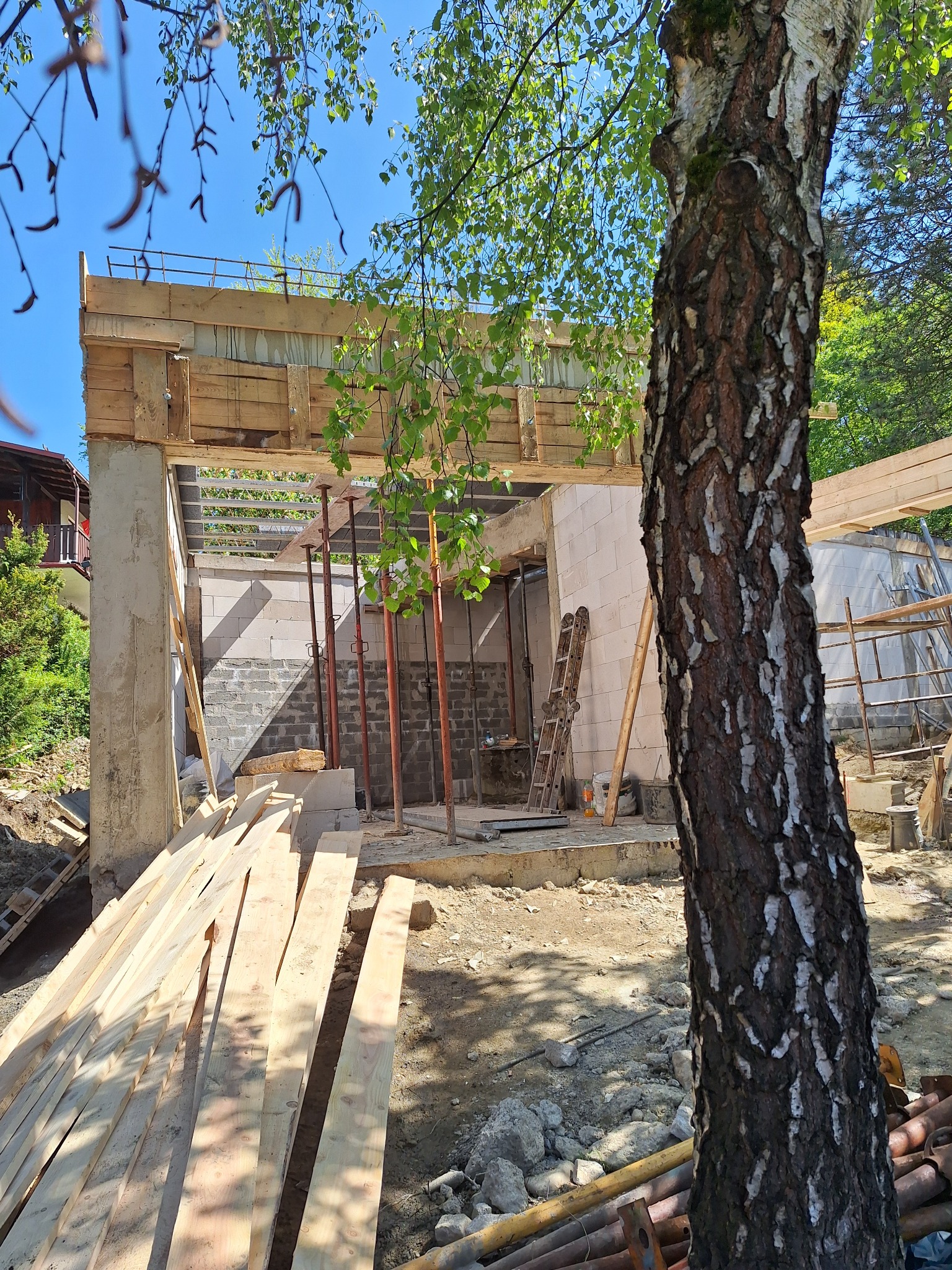 Budowa domu w trakcie realizacji: ściany z bloczków, drewniane szalunki stropu, podpory. W tle drzewo brzozy. Widok z zewnątrz na plac budowy w słoneczny dzień.