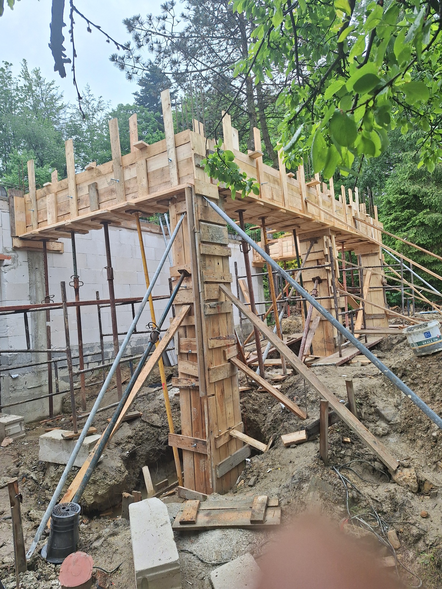 Szczegółowy widok szalunku z drewna i podpór stalowych na budowie domu, z widocznymi elementami konstrukcyjnymi i otaczającą zielenią.