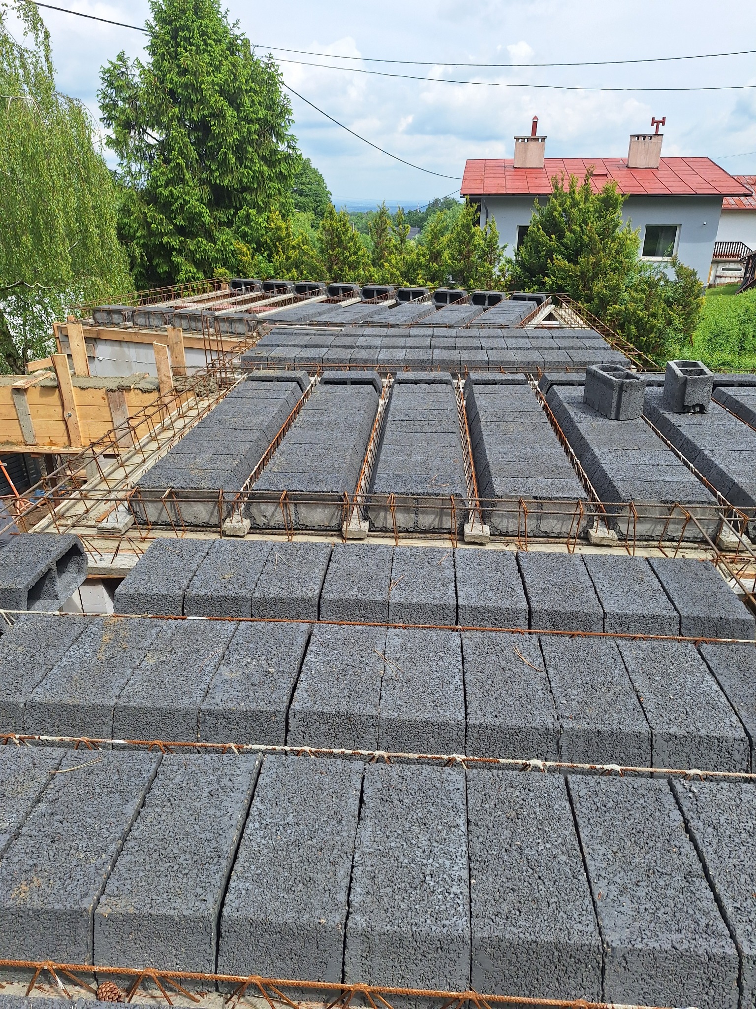 Szare bloczki stropowe ułożone na zbrojeniu, widok z góry na budowę domu jednorodzinnego w otoczeniu zieleni i zabudowań. Etap prac przed zalaniem betonem.