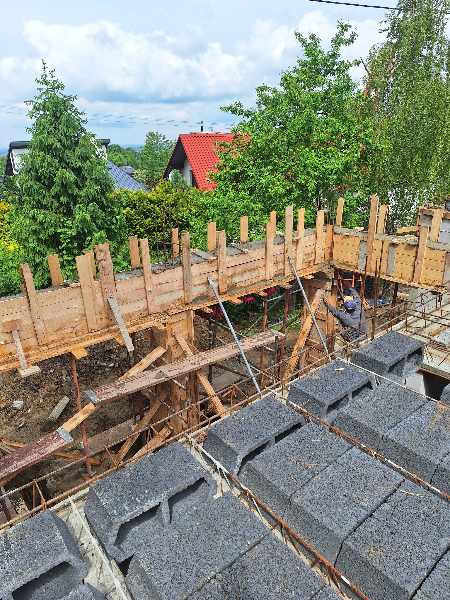 Budowa domu: szalunek drewniany, bloczki betonowe, zbrojenie. Widok z góry na plac budowy z zielenią w tle. Pracownik w kasku przy szalunku.