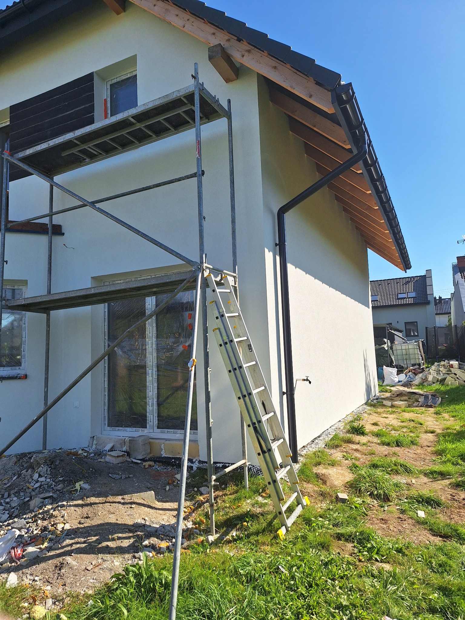 Ocieplona elewacja domu jednorodzinnego w trakcie prac wykończeniowych, widoczne rusztowanie i drabina. Okna zabezpieczone folią.