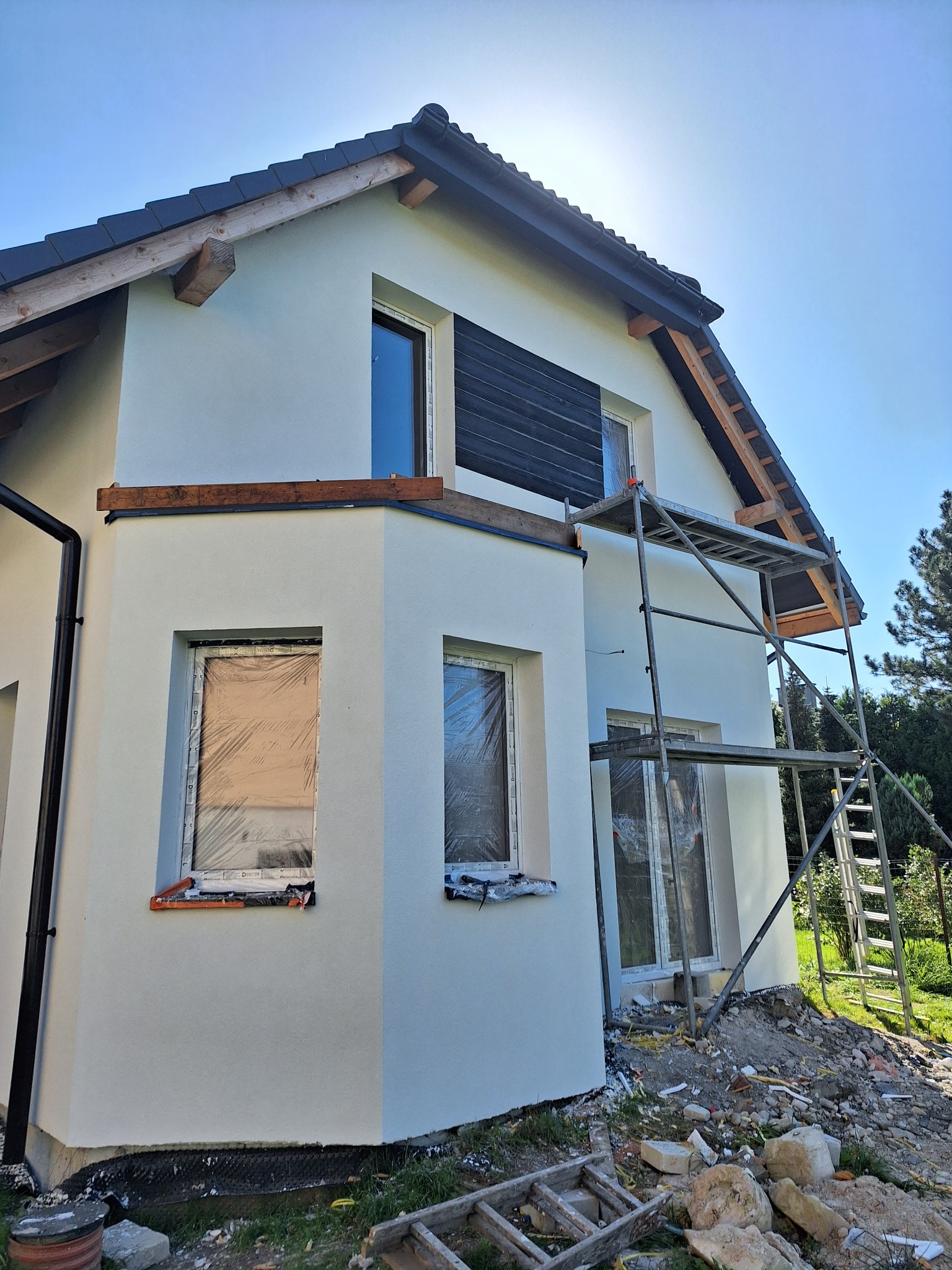 Ocieplony dom jednorodzinny z białym tynkiem, zabezpieczone okna folią, rusztowanie przy ścianie szczytowej, widoczne drewniane elementy konstrukcyjne dachu.