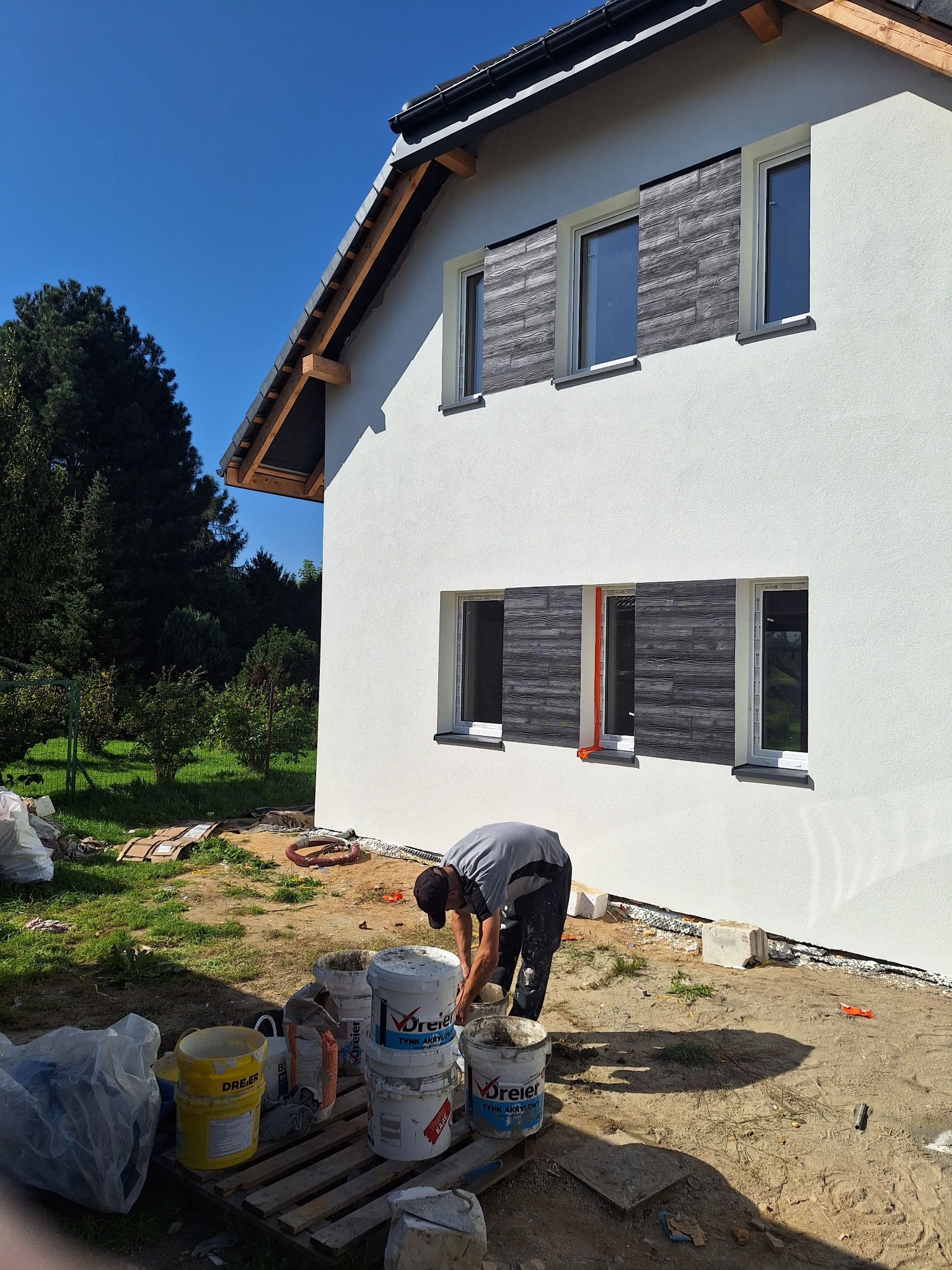 Praca przy elewacji domu: mężczyzna miesza zaprawę obok białej ściany z drewnopodobnymi panelami. Widoczne wiaderka z materiałami i narzędzia.