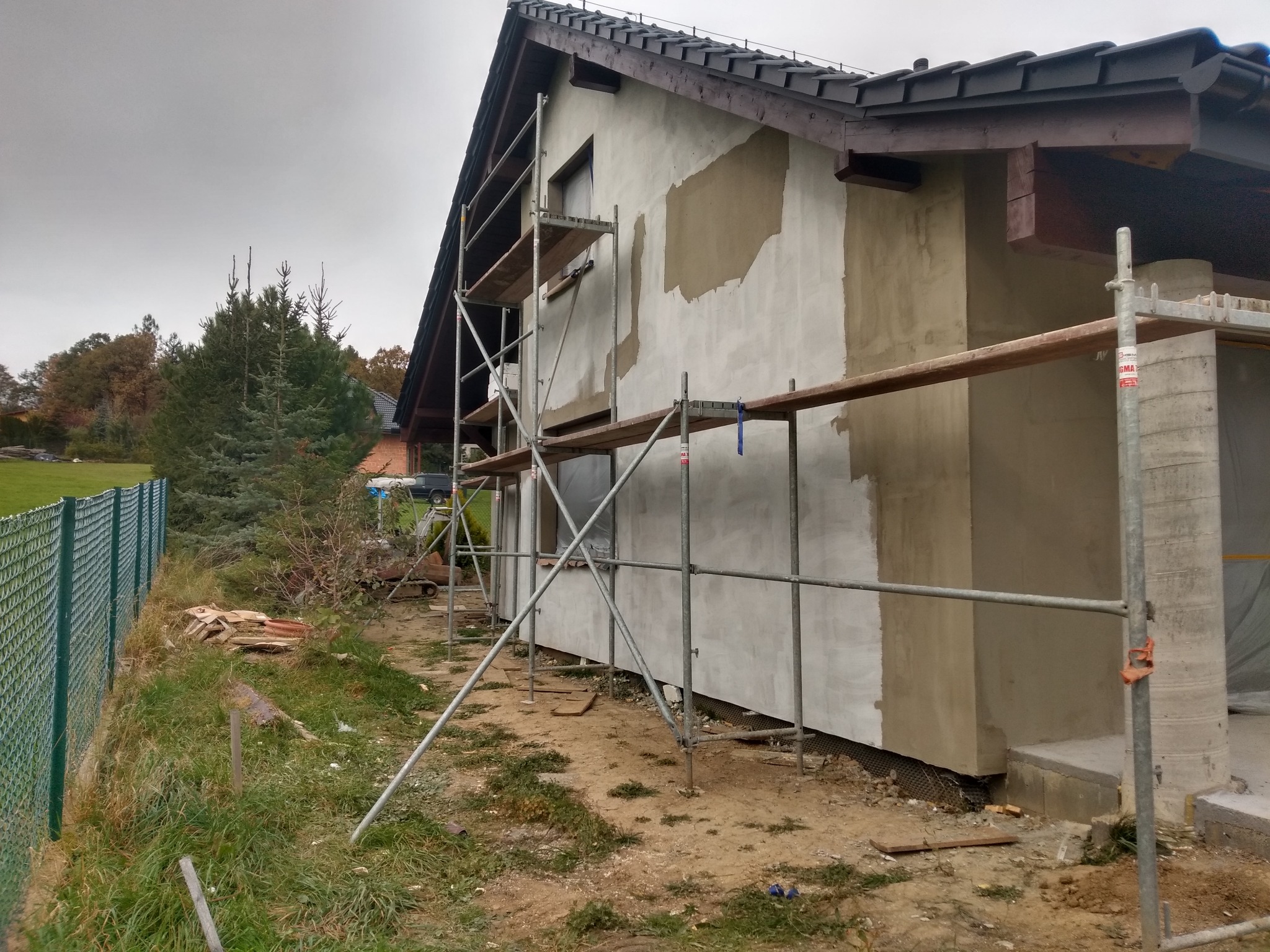 Dom w trakcie ocieplania elewacji, widoczne rusztowania i częściowo nałożony tynk. Prace w toku, widok z boku budynku, pochmurne niebo.