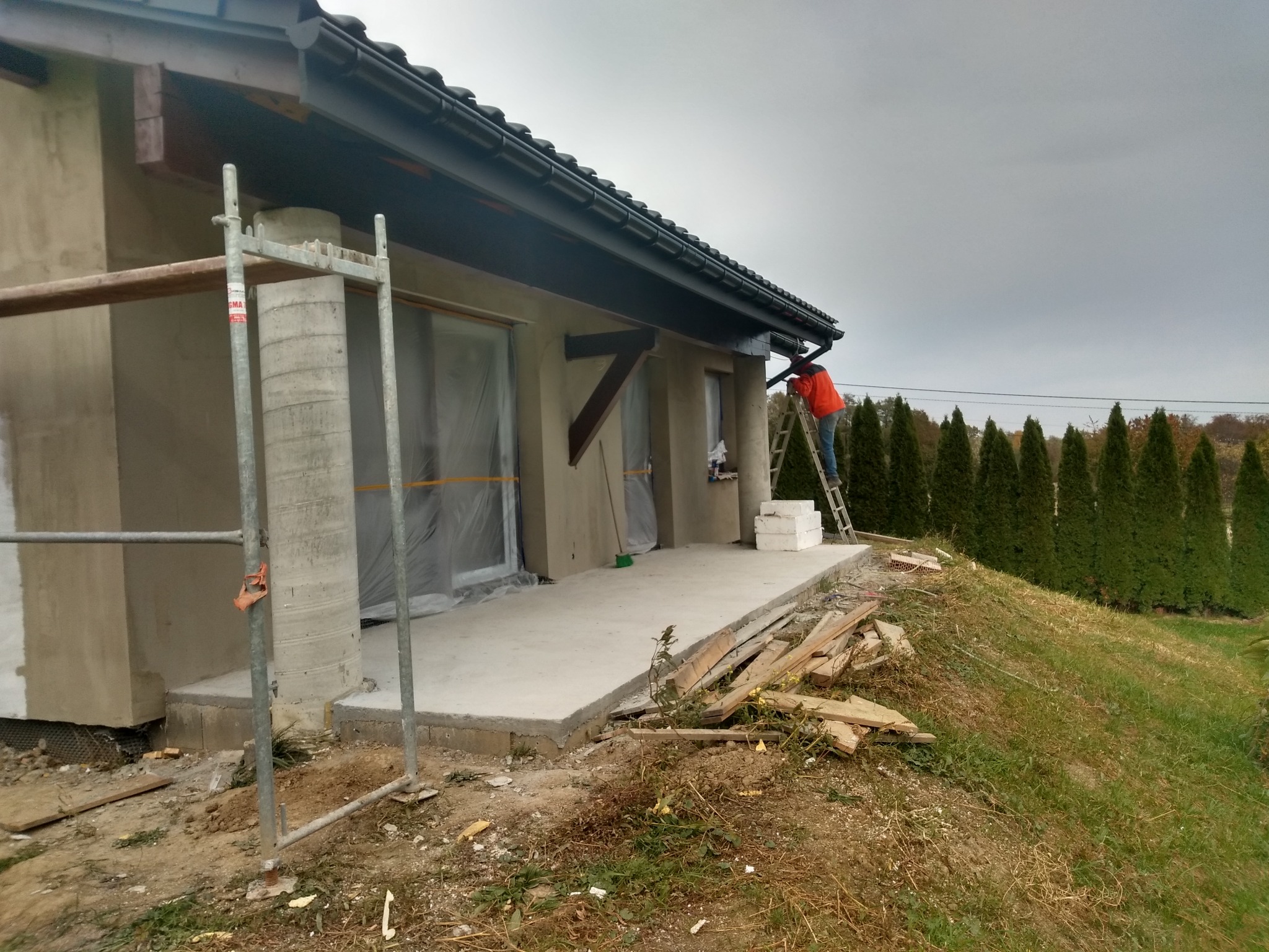 Prace wykończeniowe elewacji domu jednorodzinnego: pracownik na drabinie montuje rynnę, widoczne rusztowania i przygotowane ściany zewnętrzne.