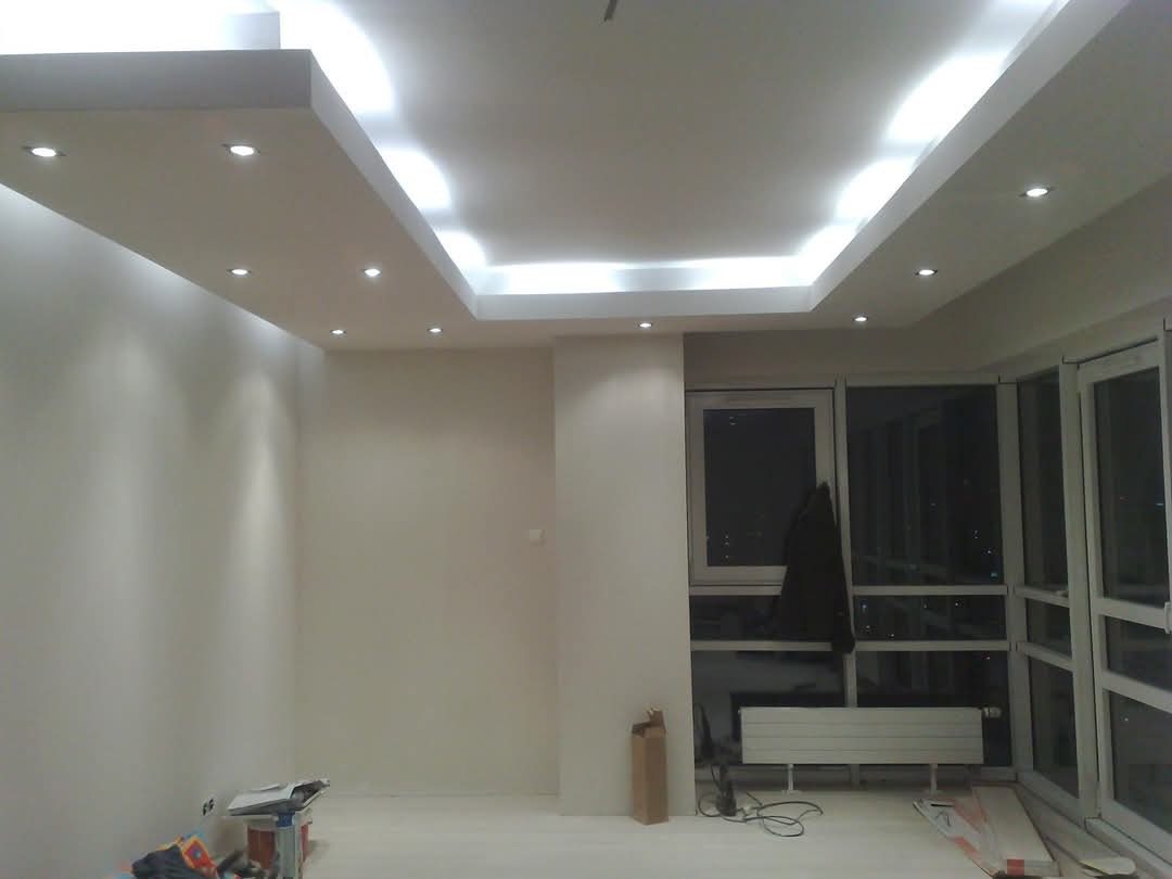 Nowoczesny sufit podwieszany z oświetleniem punktowym i ledowym w jasnym wnętrzu. Widoczne okna balkonowe i fragment podłogi z materiałami budowlanymi.