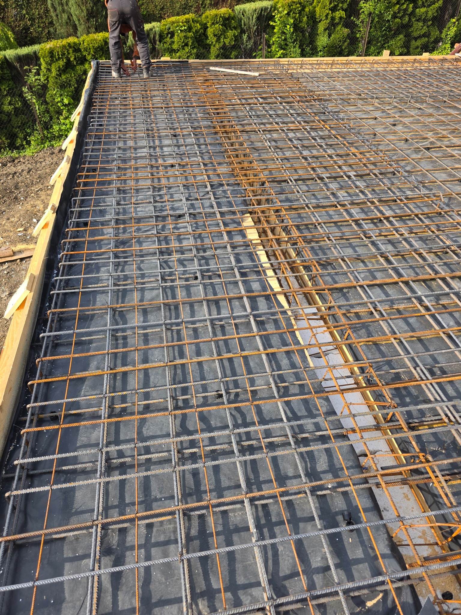 Zbrojenie płyty fundamentowej z widoczną siatką stalową i deskowaniem. Prace przygotowawcze do wylewki betonowej na czarnej folii izolacyjnej. Widok z góry.