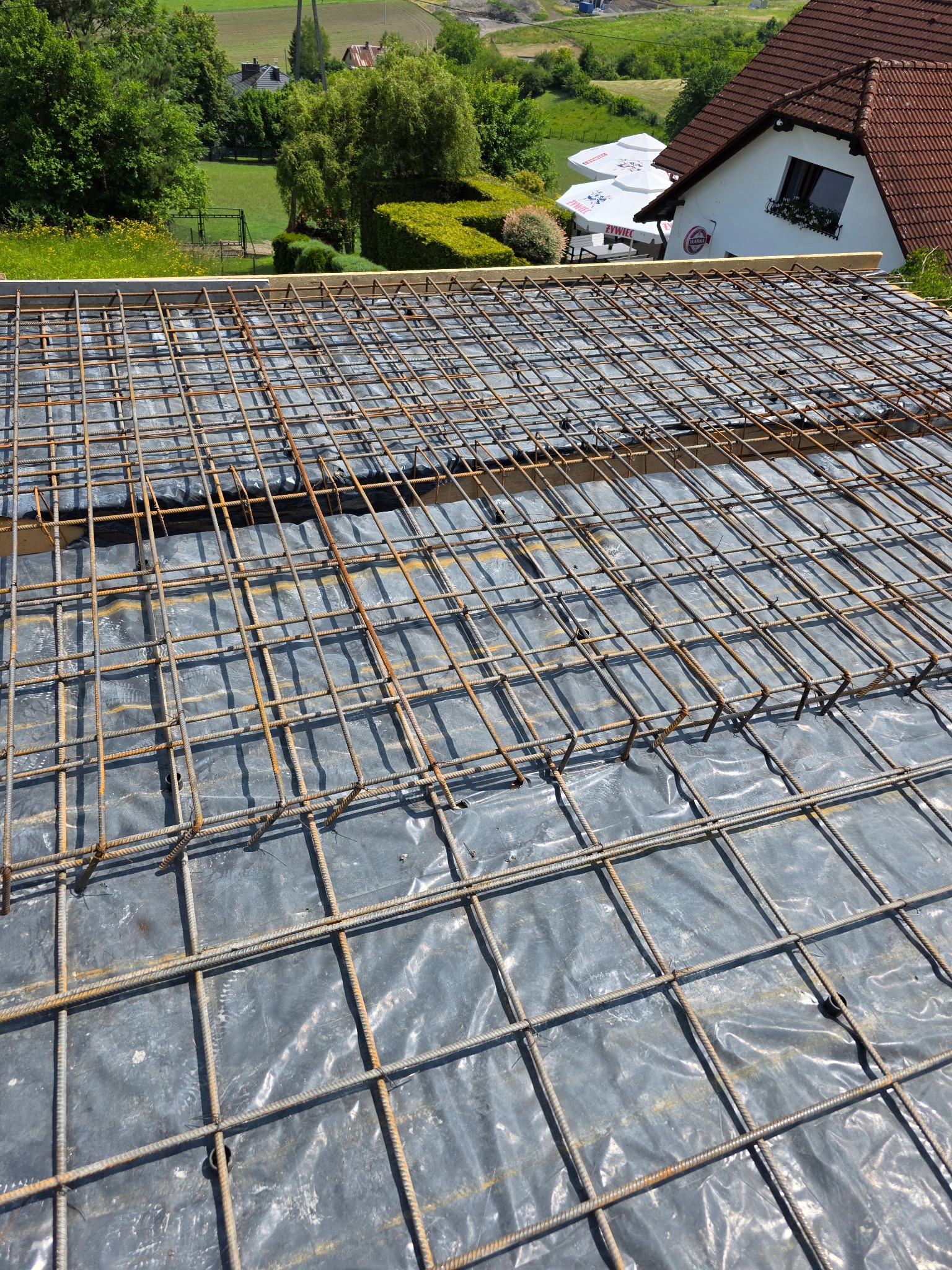 Zbrojenie stropu z siatki stalowej na folii, przygotowane do zalania betonem. W tle budynek z czerwonym dachem i zielone drzewa.