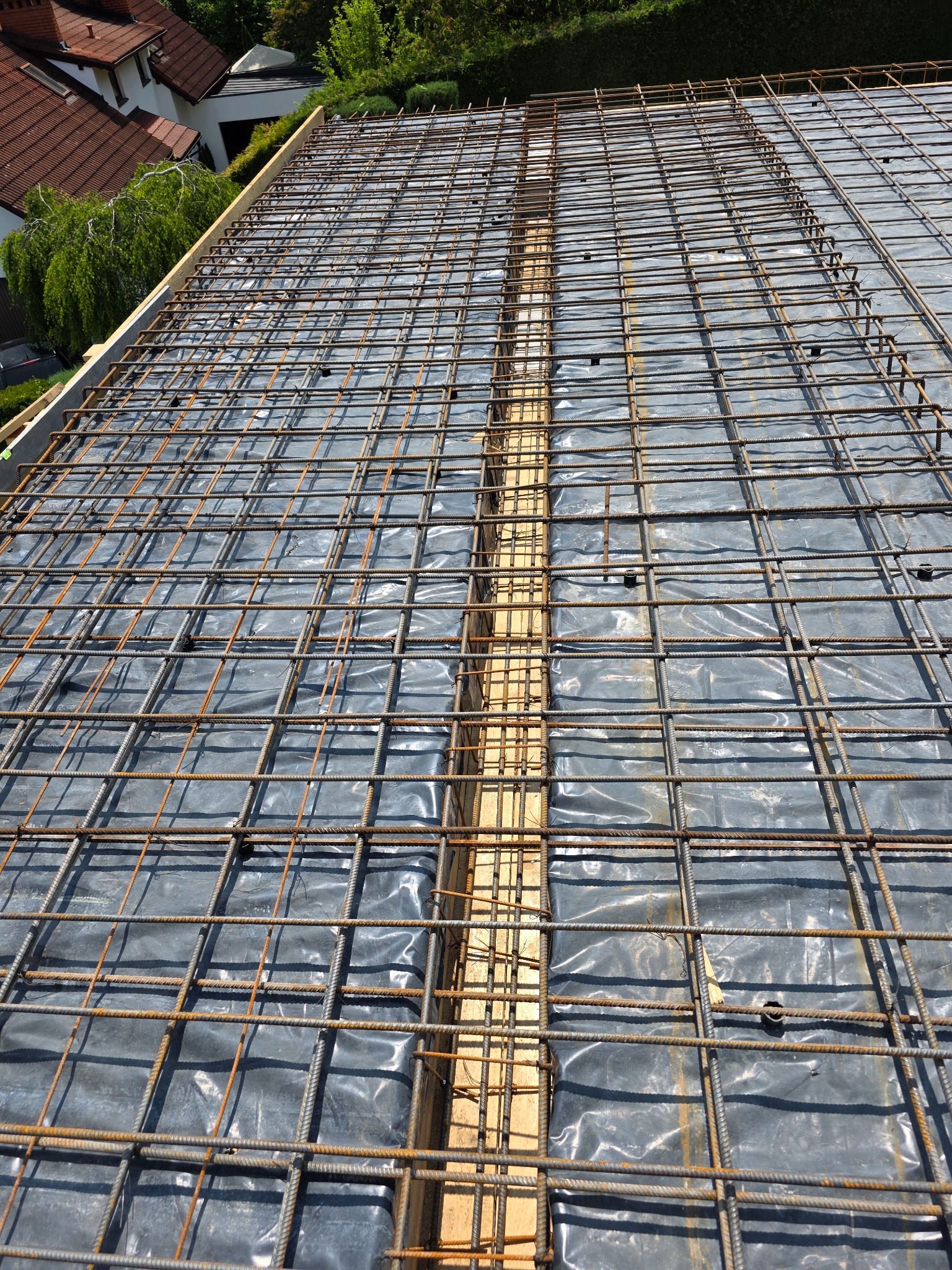 Zbrojenie stropu z widoczną folią izolacyjną i drewnianymi belkami, przygotowane do zalania betonem. Widok z góry, z elementami zabudowy w tle.