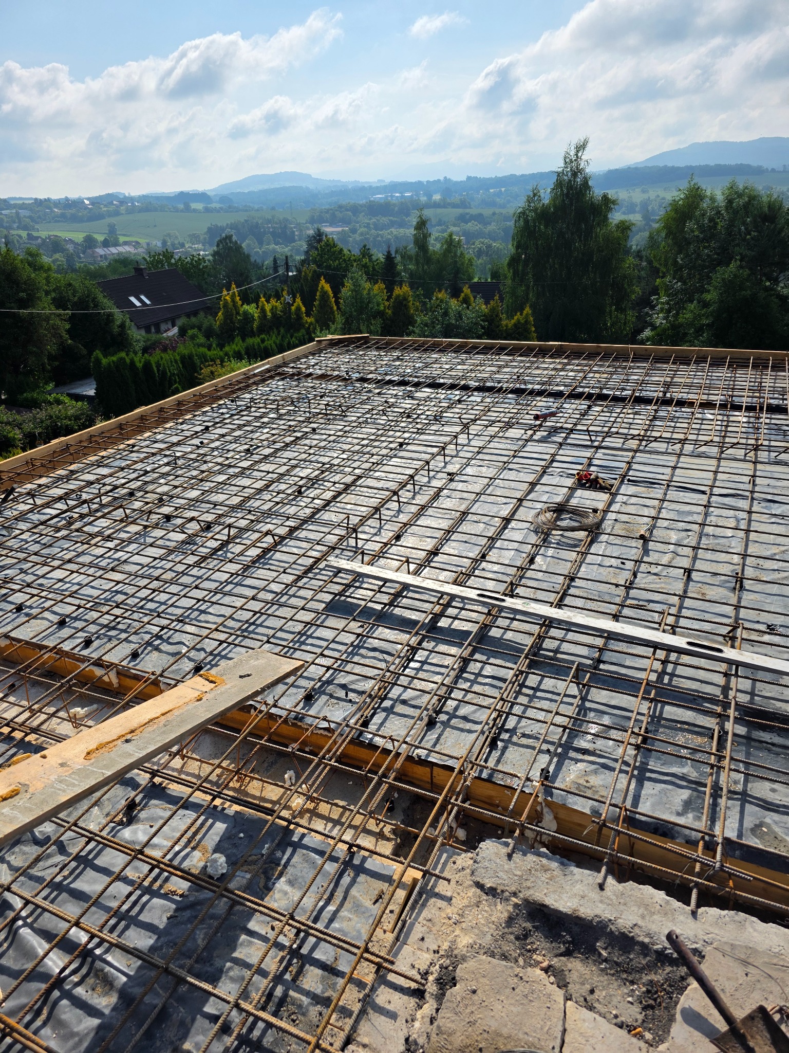Zbrojenie stropu z widokiem na pagórkowaty krajobraz. Stalowe pręty tworzą siatkę na folii, drewniane szalunki. Widoczne narzędzia i elementy montażowe.