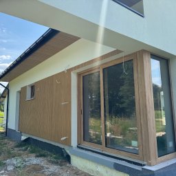 Puchalski Elewacje Łukasz Puchalski - Nowoczesny dom z elewacją z lameli w odcieniach brązu, duże okno przesuwne, widoczne elementy montażowe, częściowo otynkowana ściana, błękitne niebo.