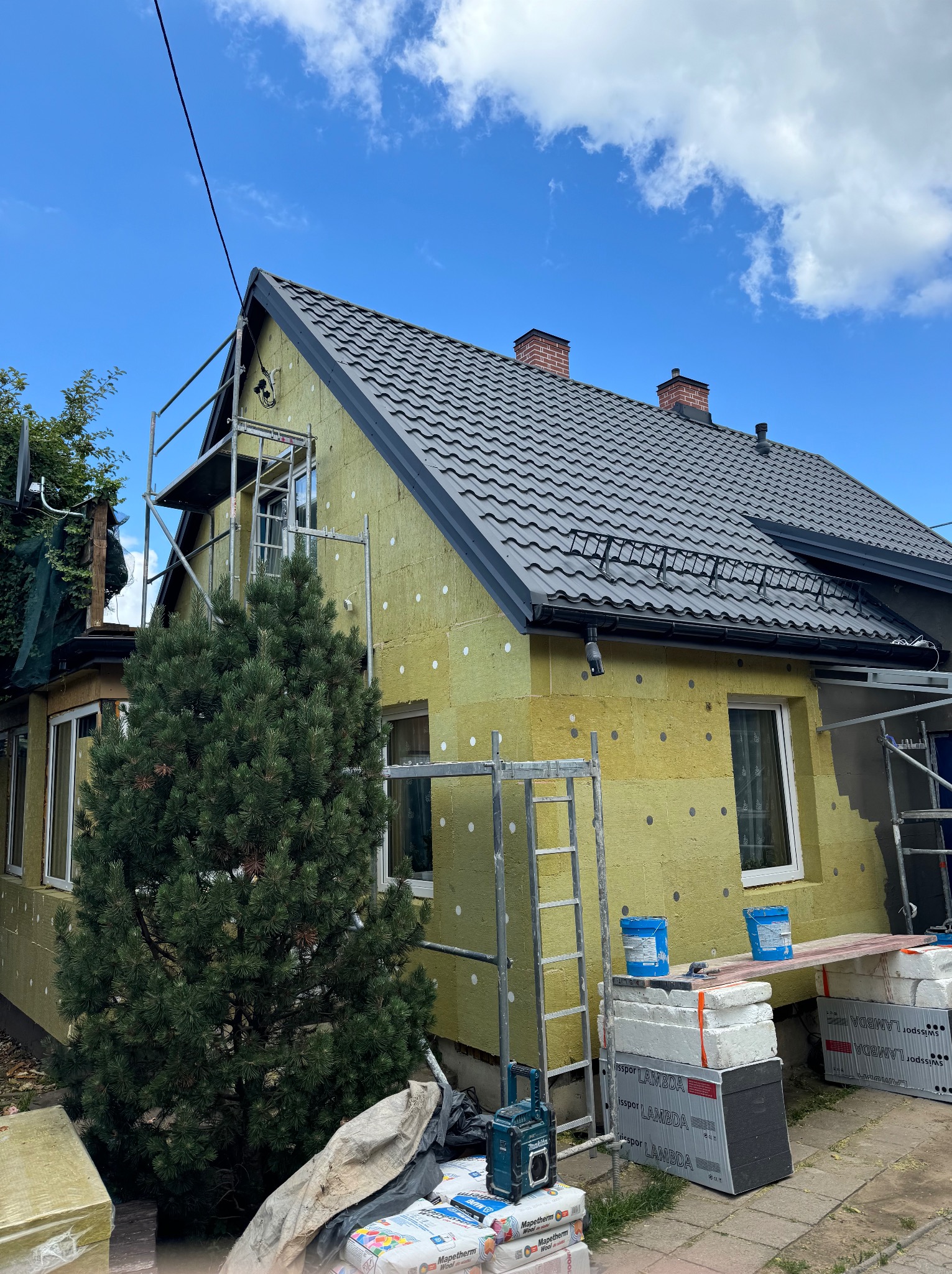 Dom w trakcie ocieplania ścian zewnętrznych, widoczne rusztowania, materiały izolacyjne i narzędzia. Prace elewacyjne w toku, zieleń w tle.