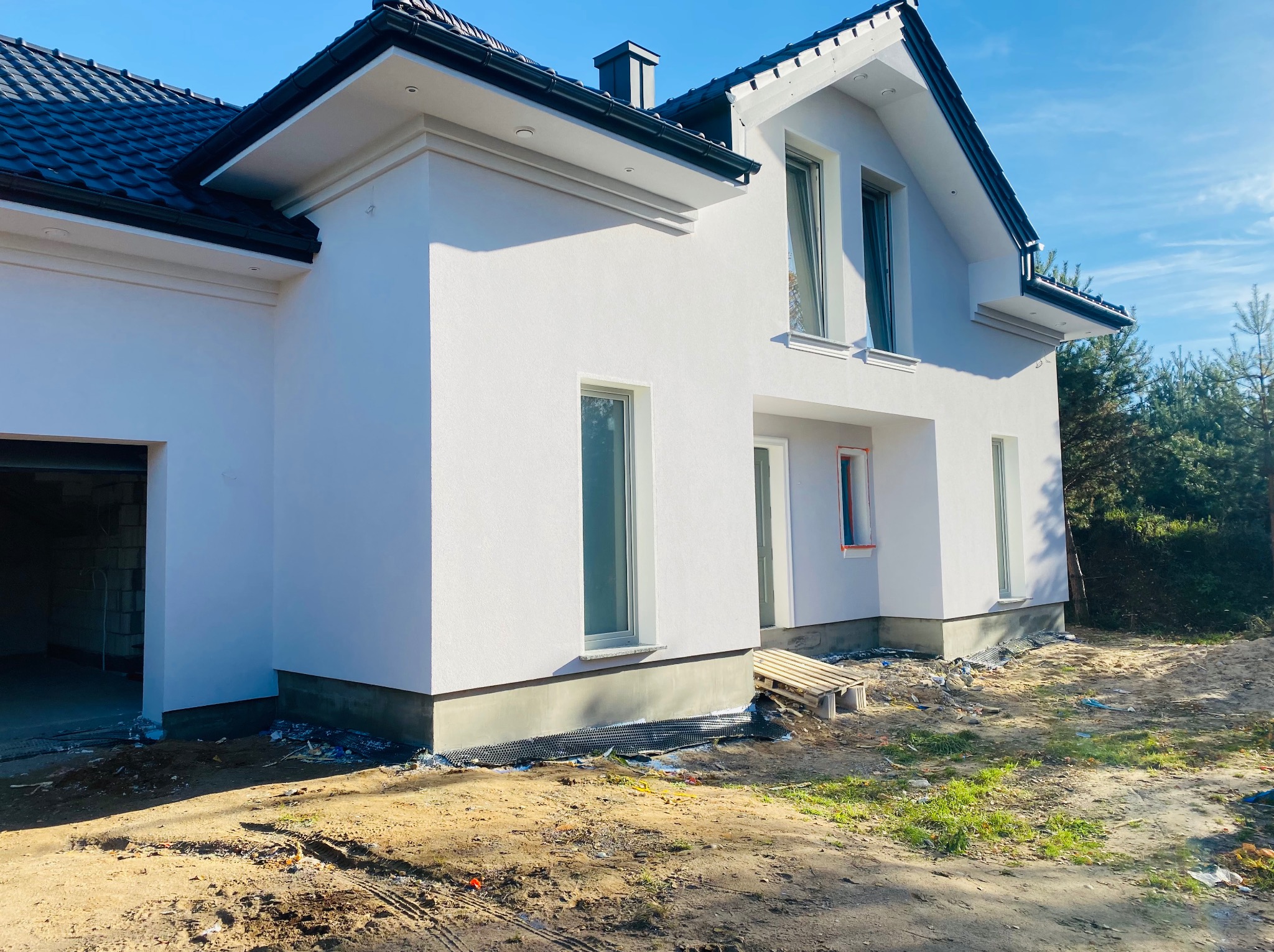 Nowoczesny, dwukondygnacyjny dom z białą elewacją i ciemną dachówką w słoneczny dzień. Widoczne detale architektoniczne i otoczenie w stanie surowym.