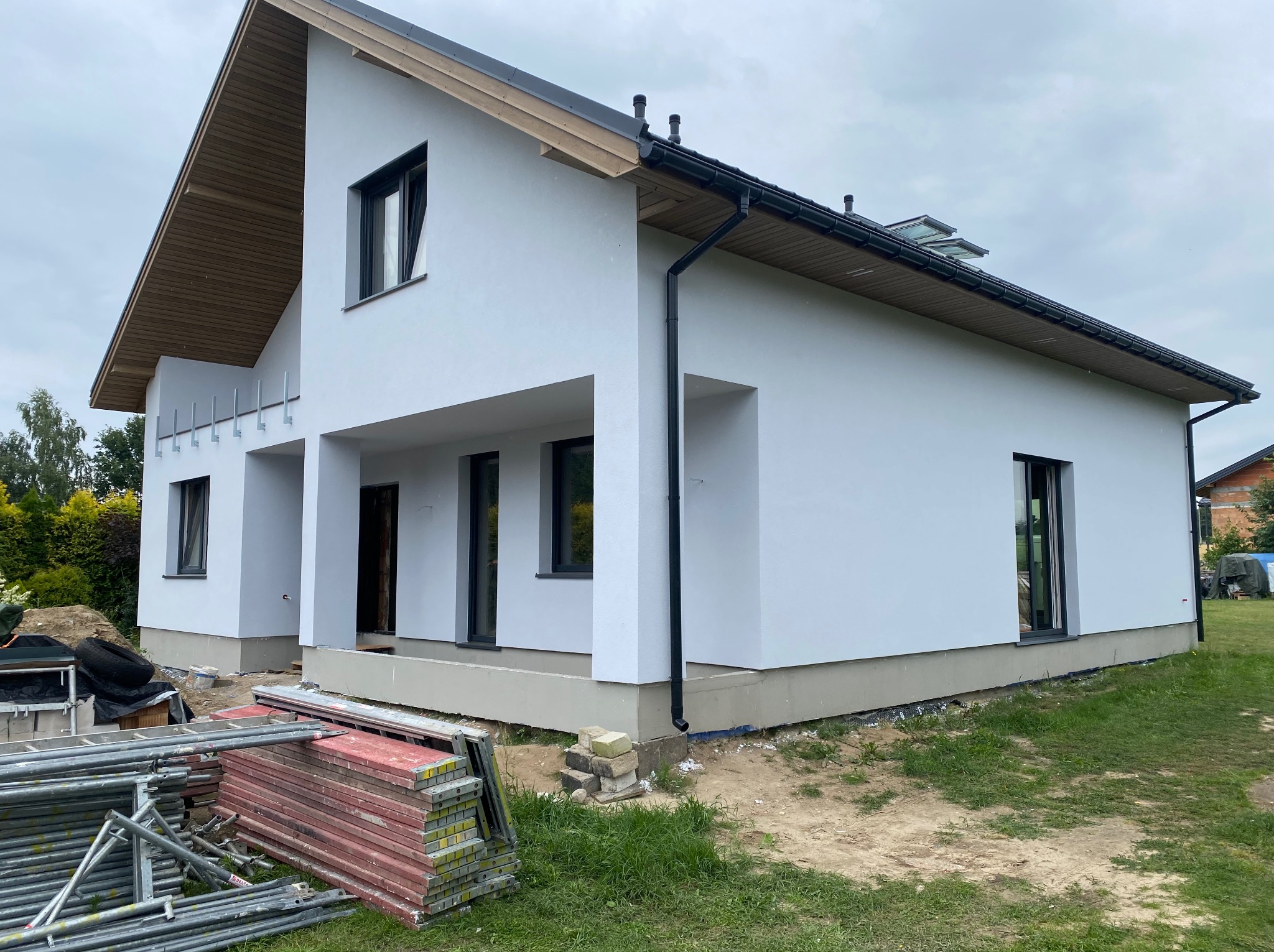 Nowoczesny, biały dom jednorodzinny z czarnymi oknami i dachem, w trakcie prac wykończeniowych. Na pierwszym planie rusztowania i materiały budowlane.