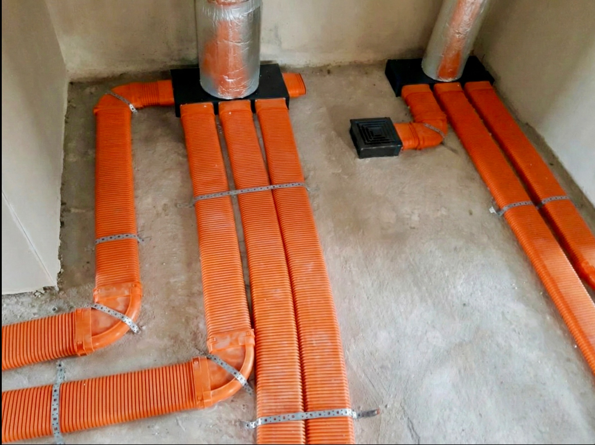 Pomarańczowe, karbowane rury wentylacyjne przymocowane metalowymi obejmami, prowadzone wzdłuż szarej, surowej podłogi i ścian. Widoczne czarne elementy montażowe.