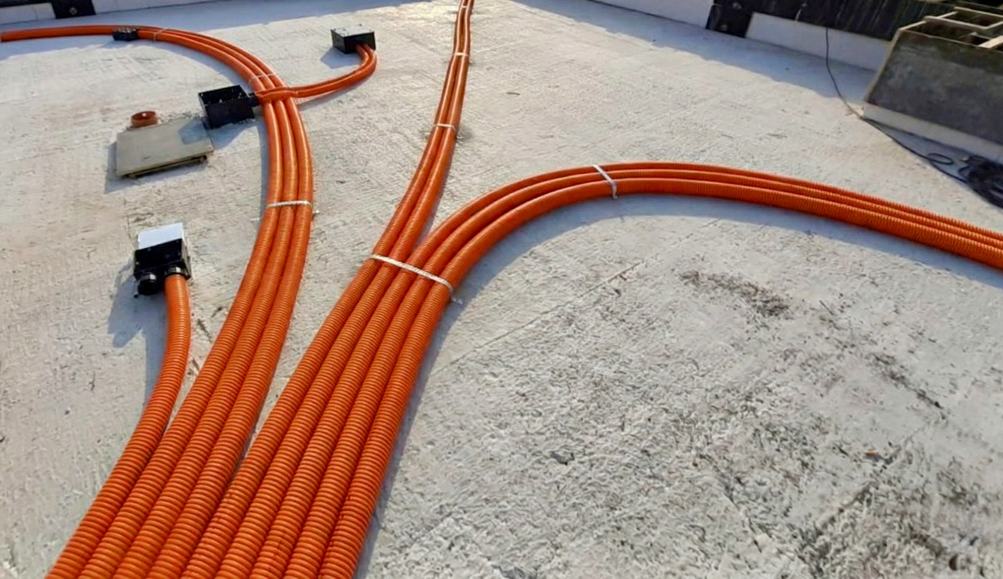 Pomarańczowe rury wentylacyjne prowadzone po szarej posadzce, połączone w wiązki białymi opaskami, z czarnymi puszkami instalacyjnymi. Instalacja rekuperacji.