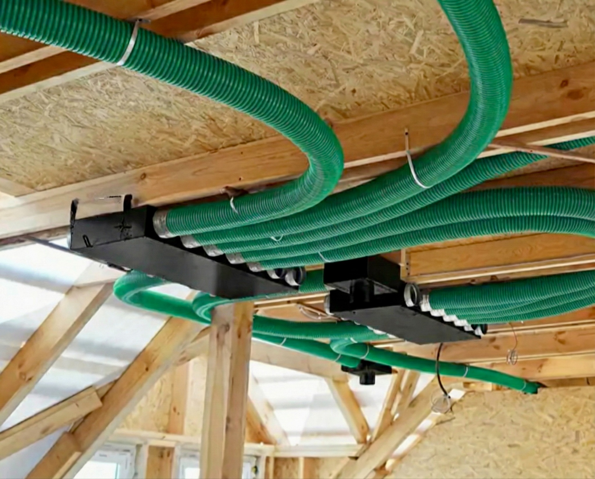 Zielone, karbowane rury wentylacyjne przymocowane do drewnianego stropu, część systemu rekuperacji w surowym stanie budynku.
