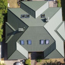 TEJADO SPÓŁKA Z OGRANICZONĄ ODPOWIEDZIALNOŚCIĄ - Widok z góry na zielony dach wielospadowy z oknami dachowymi i kominami, otoczony zielenią. Część dachu w jaśniejszym odcieniu. Widoczny fragment samochodu.