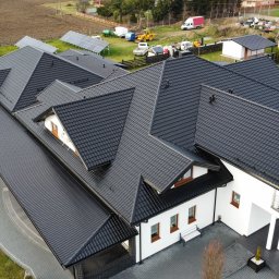 TEJADO SPÓŁKA Z OGRANICZONĄ ODPOWIEDZIALNOŚCIĄ - Czarny dach z dachówki ceramicznej na białym domu z brązowymi oknami, widok z góry. W tle panele słoneczne i sprzęt budowlany.