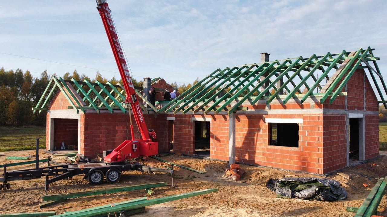 Budowa domu z czerwonej cegły, widoczna więźba dachowa z zielonych belek, na tle krajobrazu i nieba, z użyciem czerwonego dźwigu. Etap wznoszenia konstrukcji.