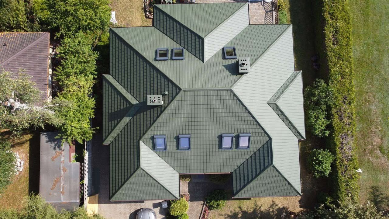 Widok z góry na zielony dach wielospadowy z oknami dachowymi i kominami, otoczony zielenią. Część dachu w jaśniejszym odcieniu. Widoczny fragment samochodu.