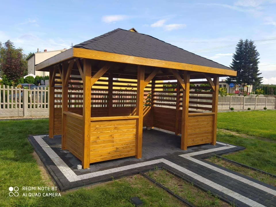 Drewniana altana z czarnym dachem z gontu bitumicznego, otoczona zielonym trawnikiem i kostką brukową. Widok na ogrodzenie i drzewa w tle.