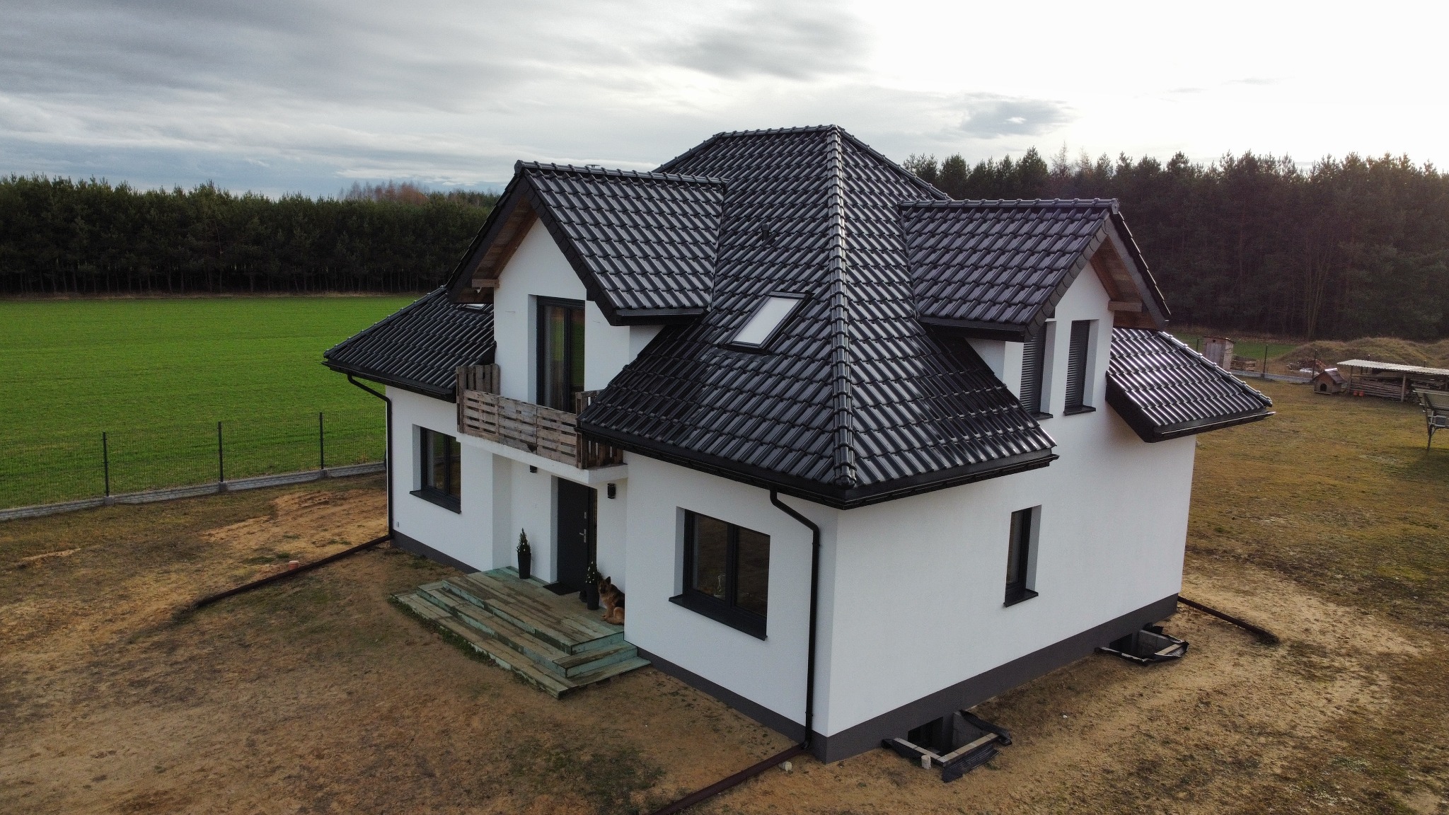 Nowoczesny dom z czarną dachówką ceramiczną i białymi ścianami, widok z góry, na tle zielonego pola i lasu. Dach z oknem połaciowym i rynnami.