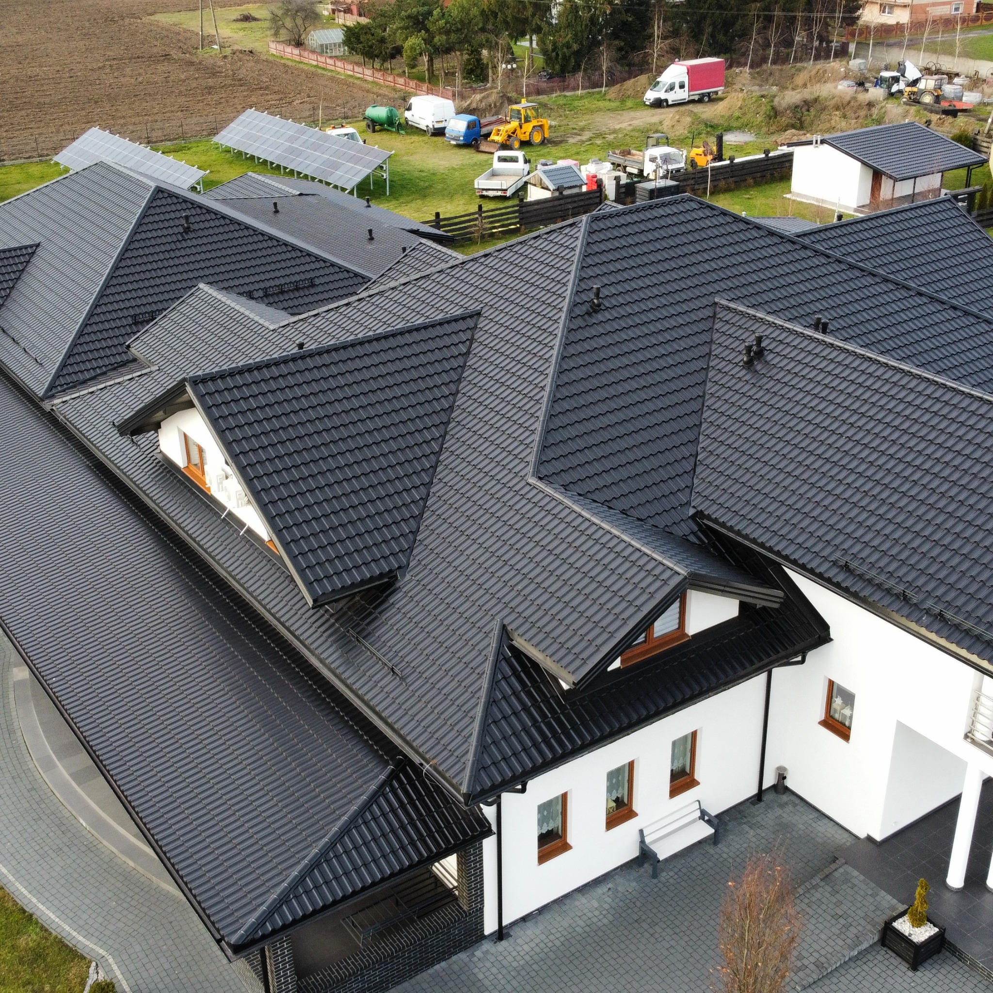 Czarny dach z dachówki ceramicznej na białym domu z brązowymi oknami, widok z góry. W tle panele słoneczne i sprzęt budowlany.