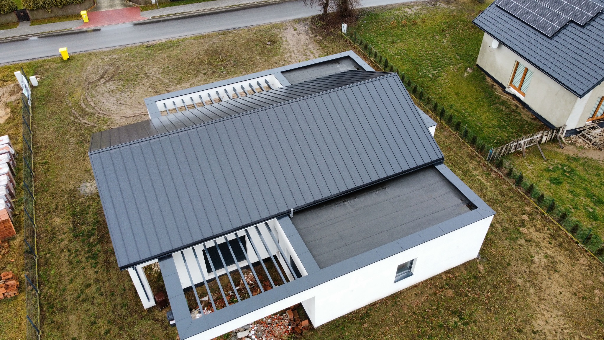 Nowoczesny dom z szarą blachą na dachu, widok z góry. Minimalistyczna architektura, płaski dach i pergola. Częściowo wykończony teren wokół budynku.