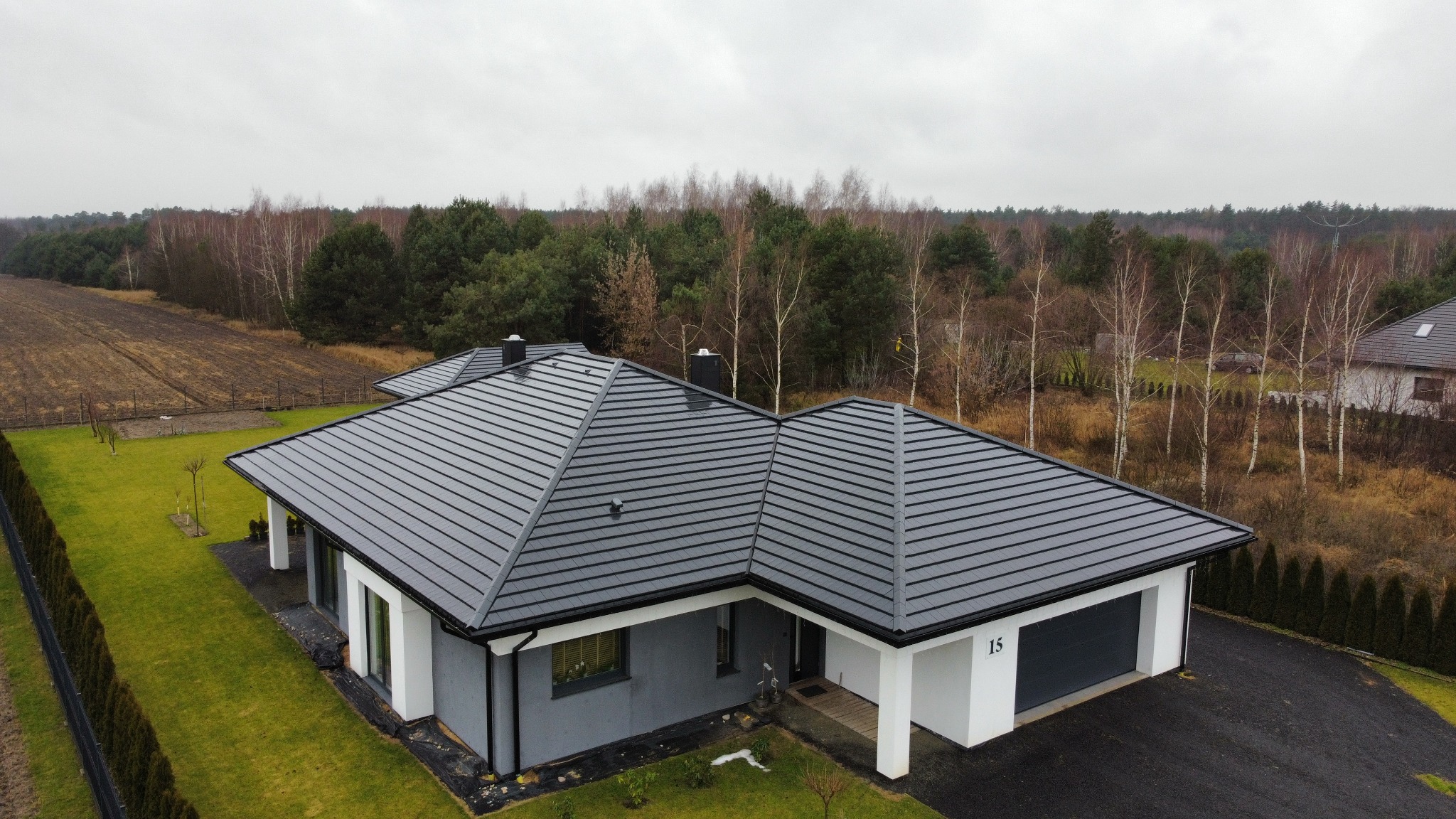 Nowoczesny dom jednorodzinny z grafitowym dachem z blachy na rąbek stojący, widok z góry, otoczony zielonym trawnikiem i drzewami. Minimalistyczna architektura.