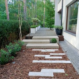 Granitex Tomasz Pietraszek - Nowoczesny taras z kamiennymi płytami ścieżki, prowadzący do domu z dużym oknem, otoczony zielenią i korą drzewną. Minimalistyczny design w naturalnym otoczeniu.