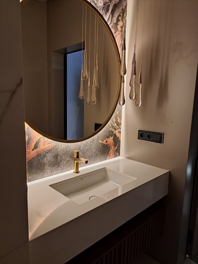 Biała, nowoczesna umywalka z blatem, złota bateria, okrągłe lustro z podświetleniem i wiszące lampy w łazience z tapetą w motywy morskie.