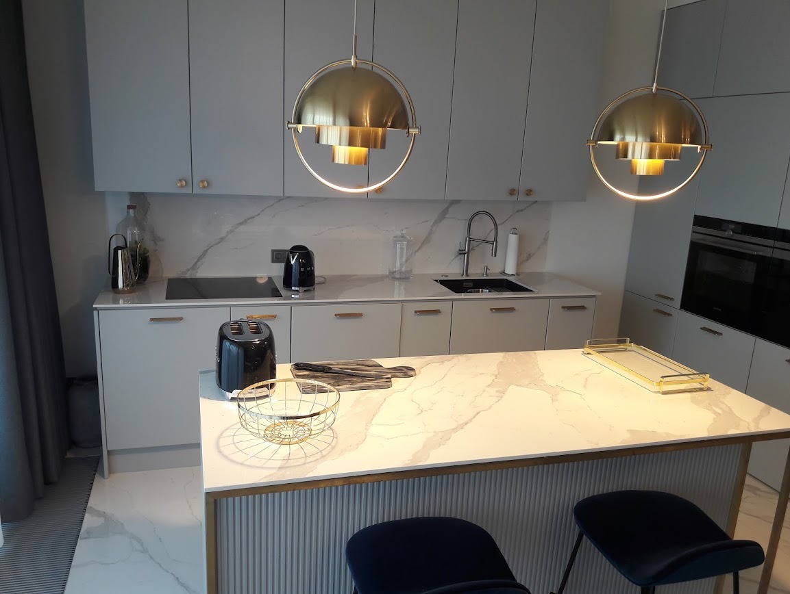 Elegancka kuchnia w szarościach z wyspą i blatem imitującym marmur. Złote lampy wiszące i dodatki podkreślają nowoczesny charakter wnętrza.