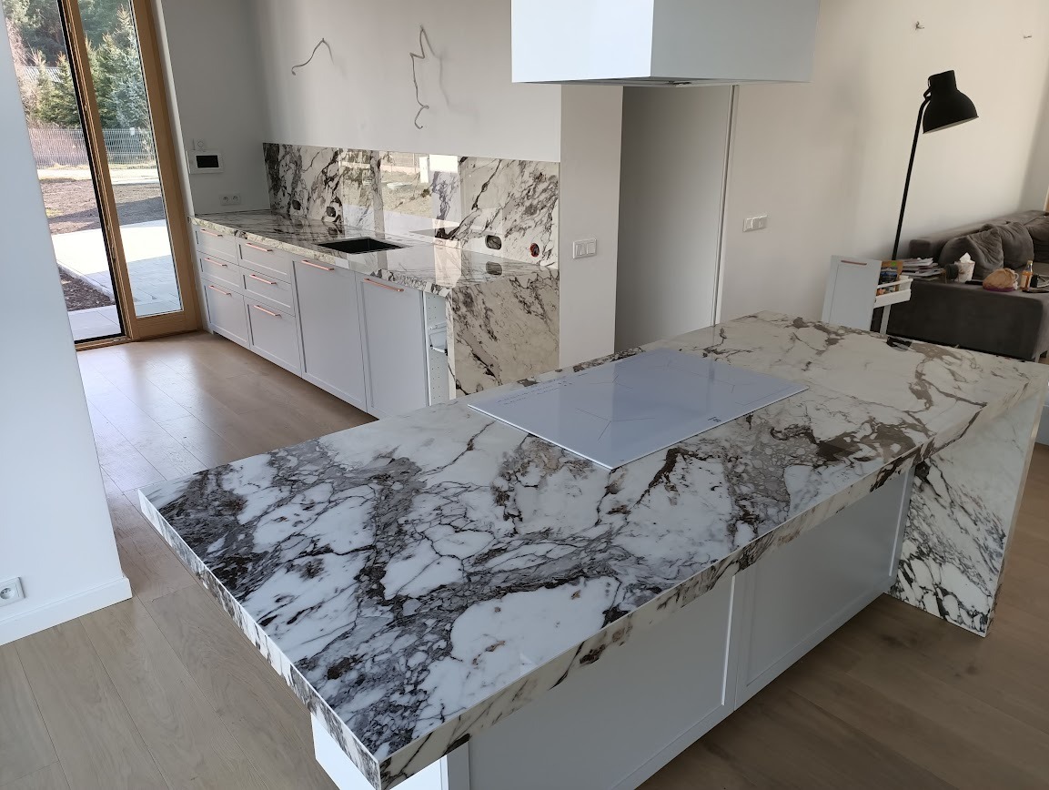 Nowoczesna kuchnia z wyspą i blatem z kamienia o unikalnym wzorze marmuru, białe szafki z miedzianymi uchwytami, płyta indukcyjna w blacie, jasne drewniane podłogi.