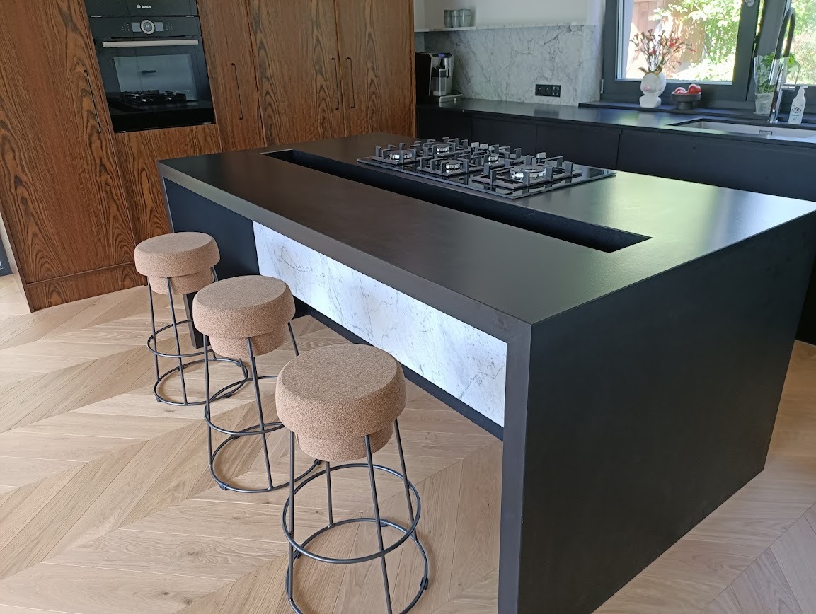 Czarny blat kuchenny z wbudowaną płytą grzewczą i marmurowym wykończeniem, uzupełniony o trzy korkowe hokery na metalowych nogach, na tle parkietu w jodełkę.