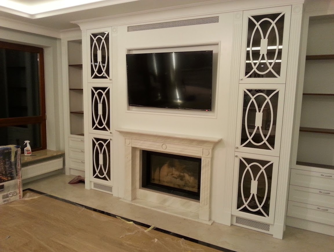 Elegancki salon z kominkiem obudowanym jasnym kamieniem, nad którym wisi telewizor. Po bokach symetryczne, białe szafki z dekoracyjnymi, ażurowymi frontami. Klasyczny design.