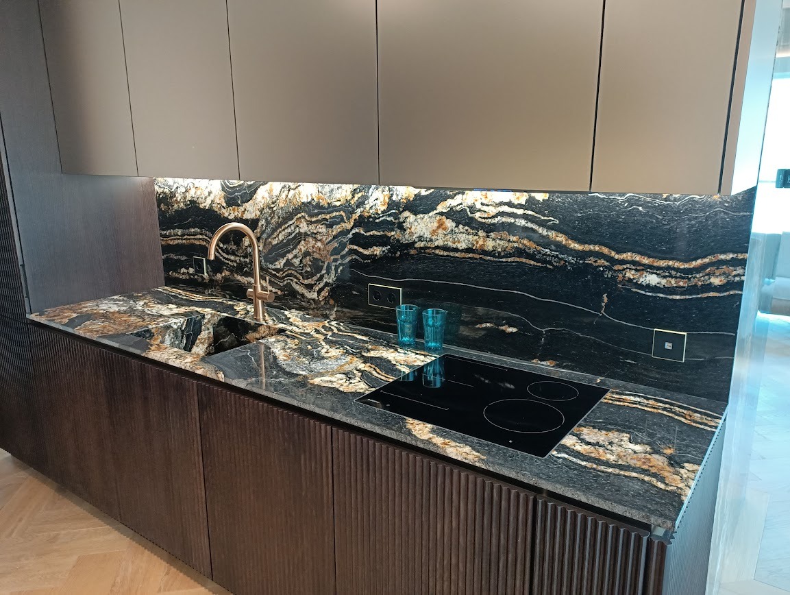 Ekskluzywny blat kamienny z unikalnym wzorem żyłkowania, dopasowany do ściany nad blatem. Złota bateria i płyta indukcyjna wkomponowane w całość. Dwa turkusowe szkła.