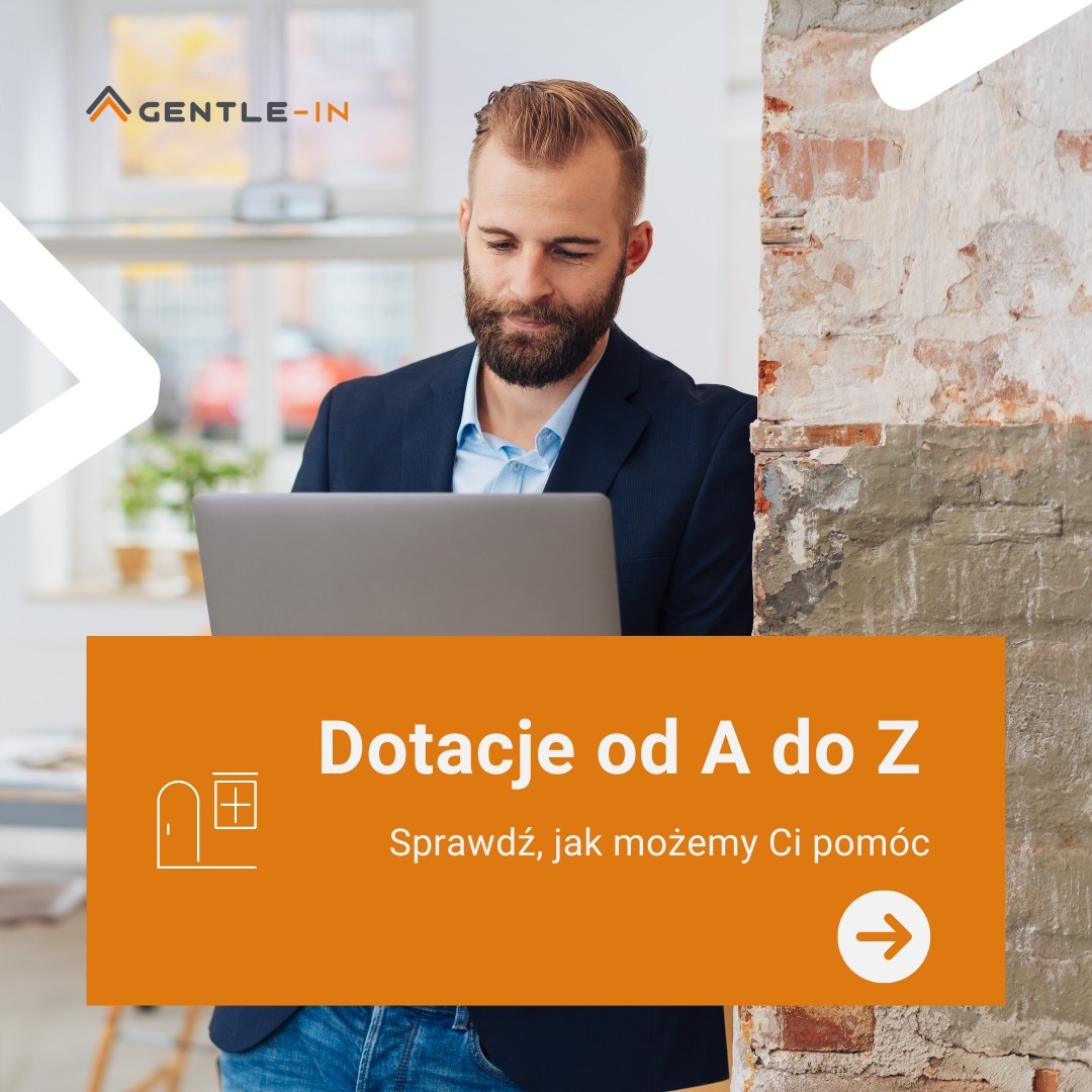 Mężczyzna z brodą, w marynarce, pracuje na laptopie. Obok niego fragment ceglanej ściany. Na pierwszym planie pomarańczowy baner z napisem 'Dotacje od A do Z'.