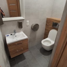 BJP DEVELOPMENT INVESTMENT SPÓŁKA Z OGRANICZONĄ ODPOWIEDZIALNOŚCIĄ - Wykończona łazienka w szarościach z drewnianymi akcentami: umywalka z szafką, toaleta podwieszana i lustro w białej ramie. Widoczny dozownik mydła i podajnik ręczników.