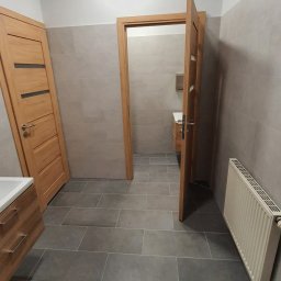 BJP DEVELOPMENT INVESTMENT SPÓŁKA Z OGRANICZONĄ ODPOWIEDZIALNOŚCIĄ - Wykończona łazienka z szarą płytką podłogową, drewnianymi drzwiami i grzejnikiem. Widok na umywalkę w głębi. Częściowo widoczny sufit podwieszany.