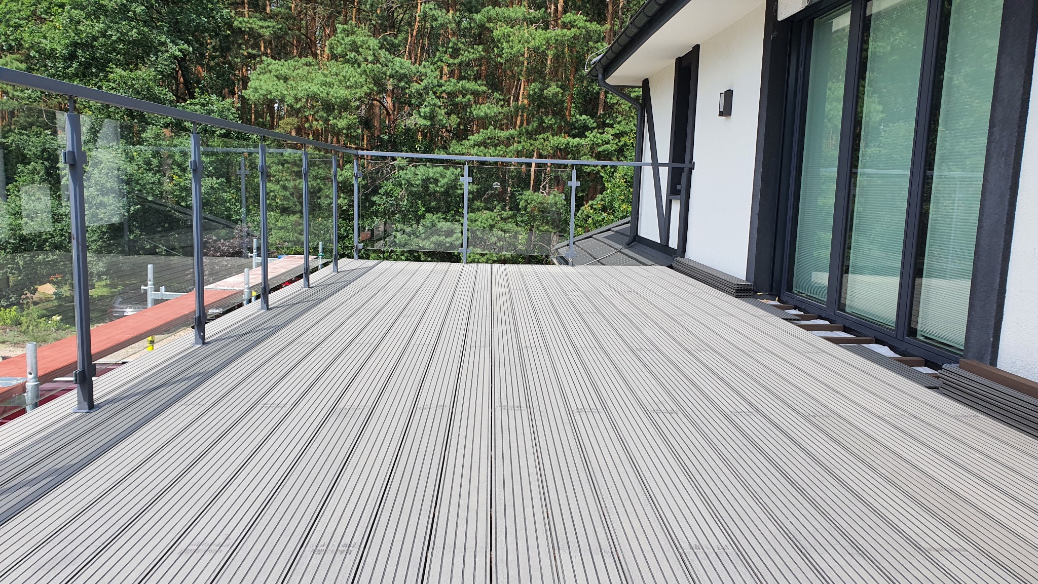 Nowoczesny balkon z szarą deską kompozytową i szklaną balustradą na tle zielonego lasu. Widoczne elementy montażowe i fragment elewacji budynku.