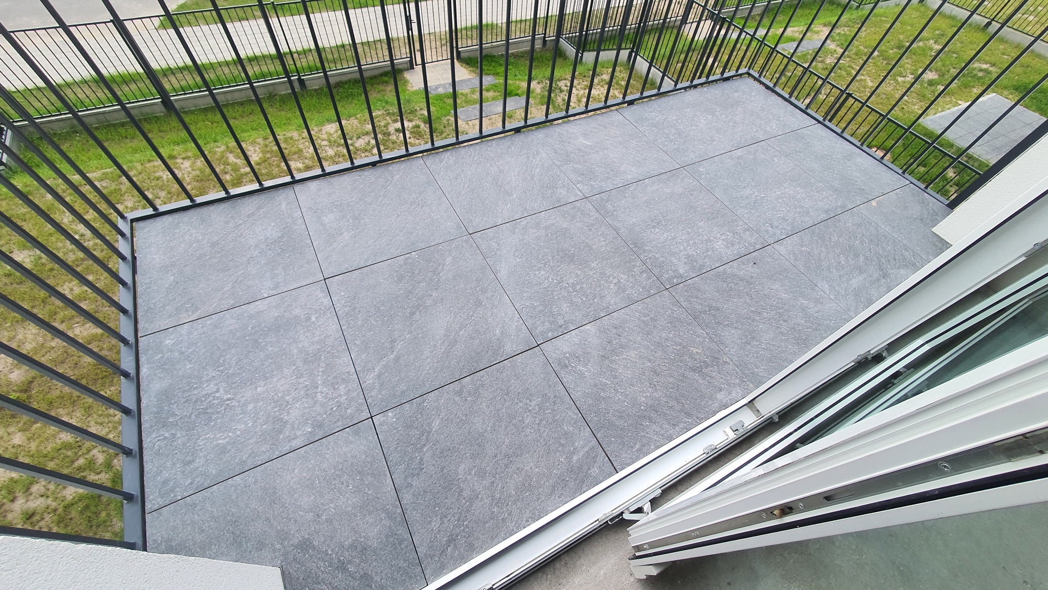 Wykończony balkon z szarą, kwadratową terakotą i czarną, metalową balustradą. Widok z góry na podłogę balkonu i otwarte okno balkonowe.